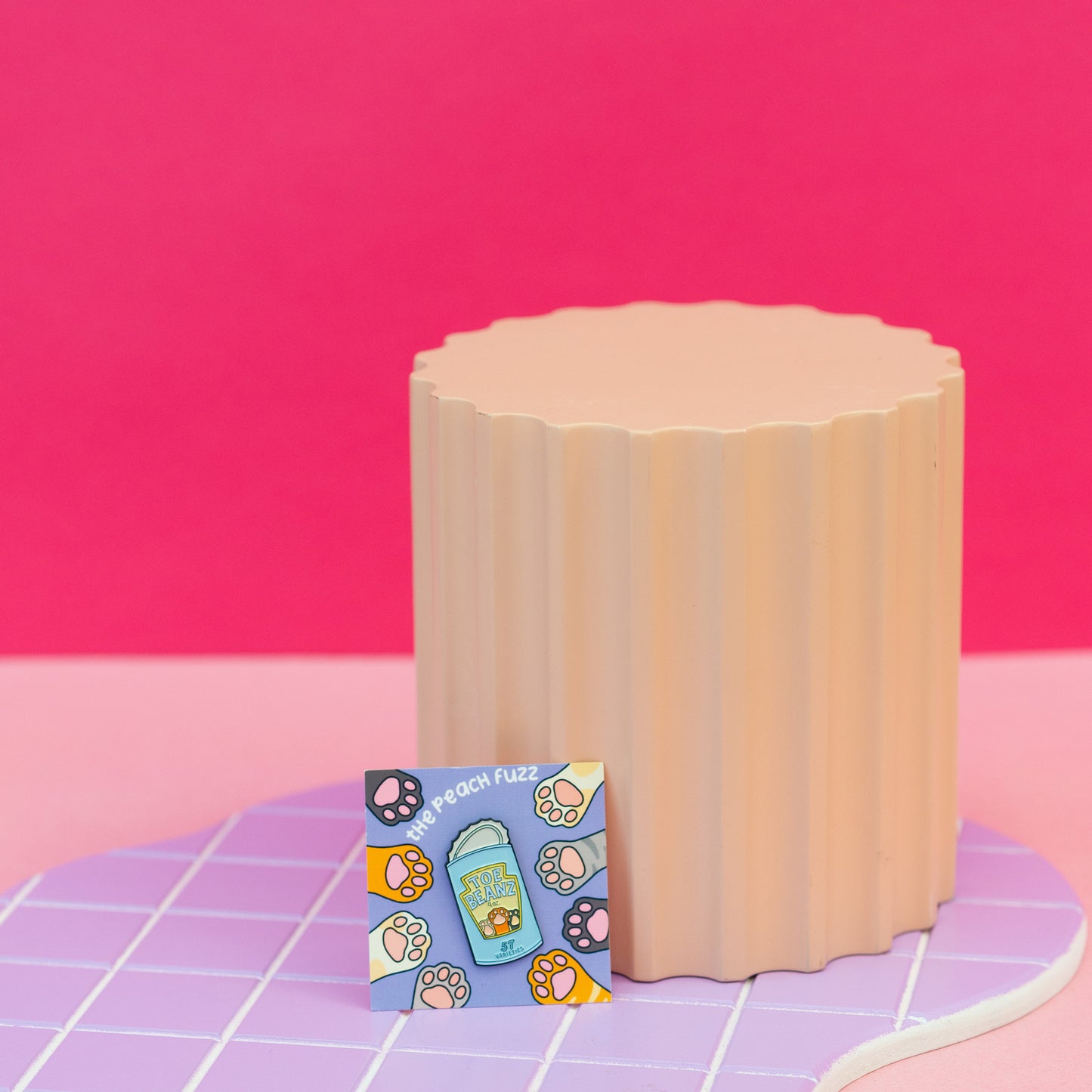 A soft enamel pin of a can of beans with the lid open and the label for the beans says “toe beans” with three little cat paws reaching up. The backing card has dark brown, yellow, white, orange, and gray cat paws reaching towards the center where the pin is.