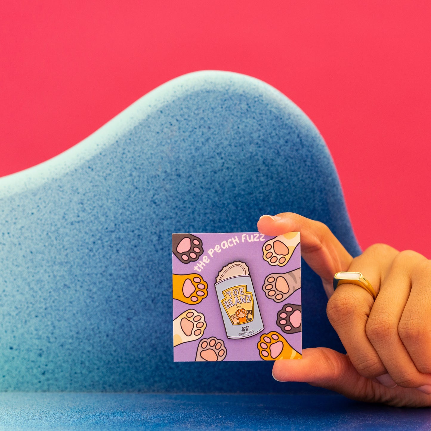 A soft enamel pin of a can of beans with the lid open and the label for the beans says “toe beans” with three little cat paws reaching up. The backing card has dark brown, yellow, white, orange, and gray cat paws reaching towards the center where the pin is.