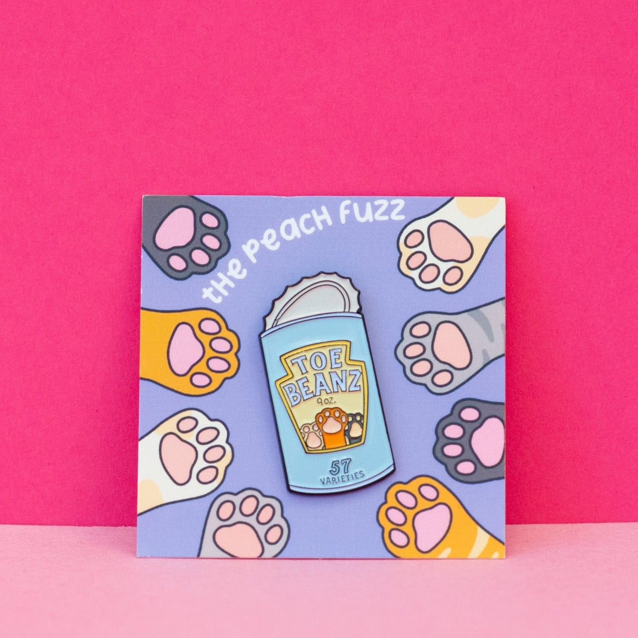A soft enamel pin of a can of beans with the lid open and the label for the beans says “toe beans” with three little cat paws reaching up. The backing card has dark brown, yellow, white, orange, and gray cat paws reaching towards the center where the pin is.