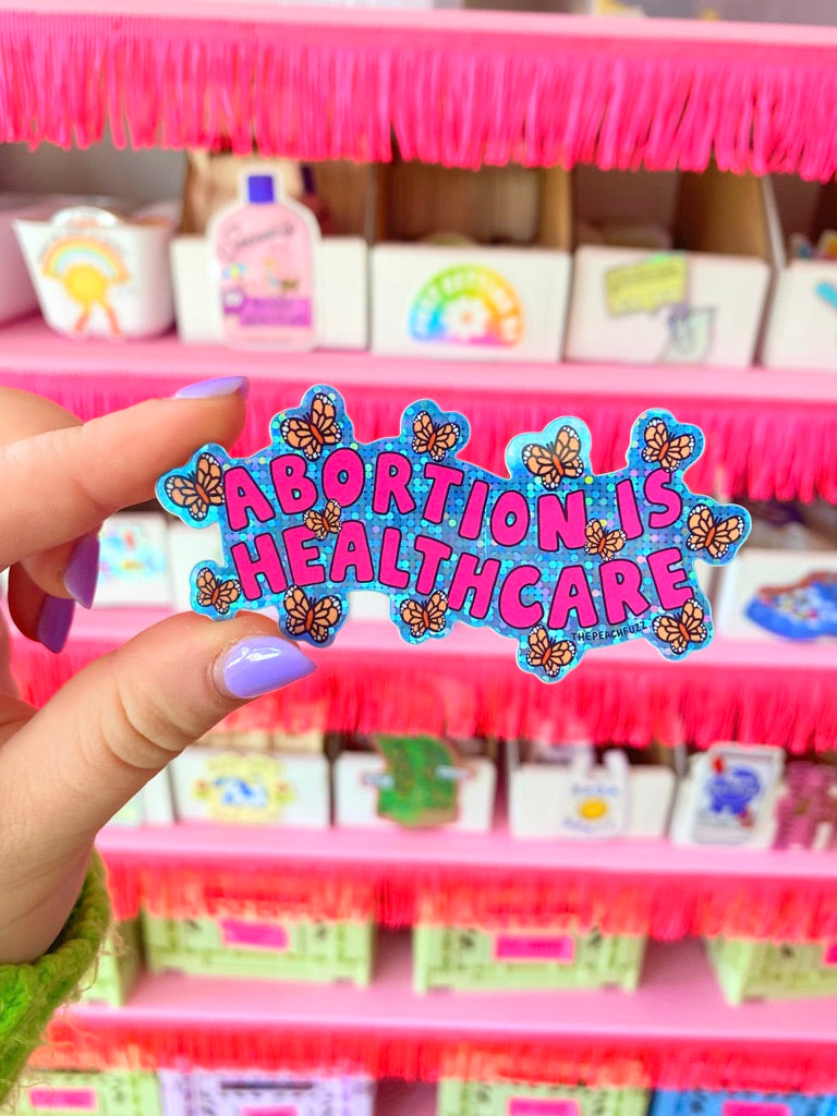 “Abortion Is Healthcare” Vinyl sticker with glossy finish and glitter detail. Measures 3.3 x 1.7 inches. - The Peach Fuzz