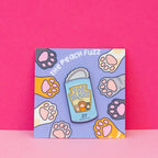 A soft enamel pin of a can of beans with the lid open and the label for the beans says “toe beans” with three little cat paws reaching up. The backing card has dark brown, yellow, white, orange, and gray cat paws reaching towards the center where the pin is.