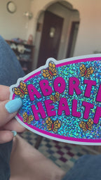 A vinyl bumper sticker with glitter detail, featuring butterflies surrounding the text 'Abortion is Healthcare’. - The Peach Fuzz