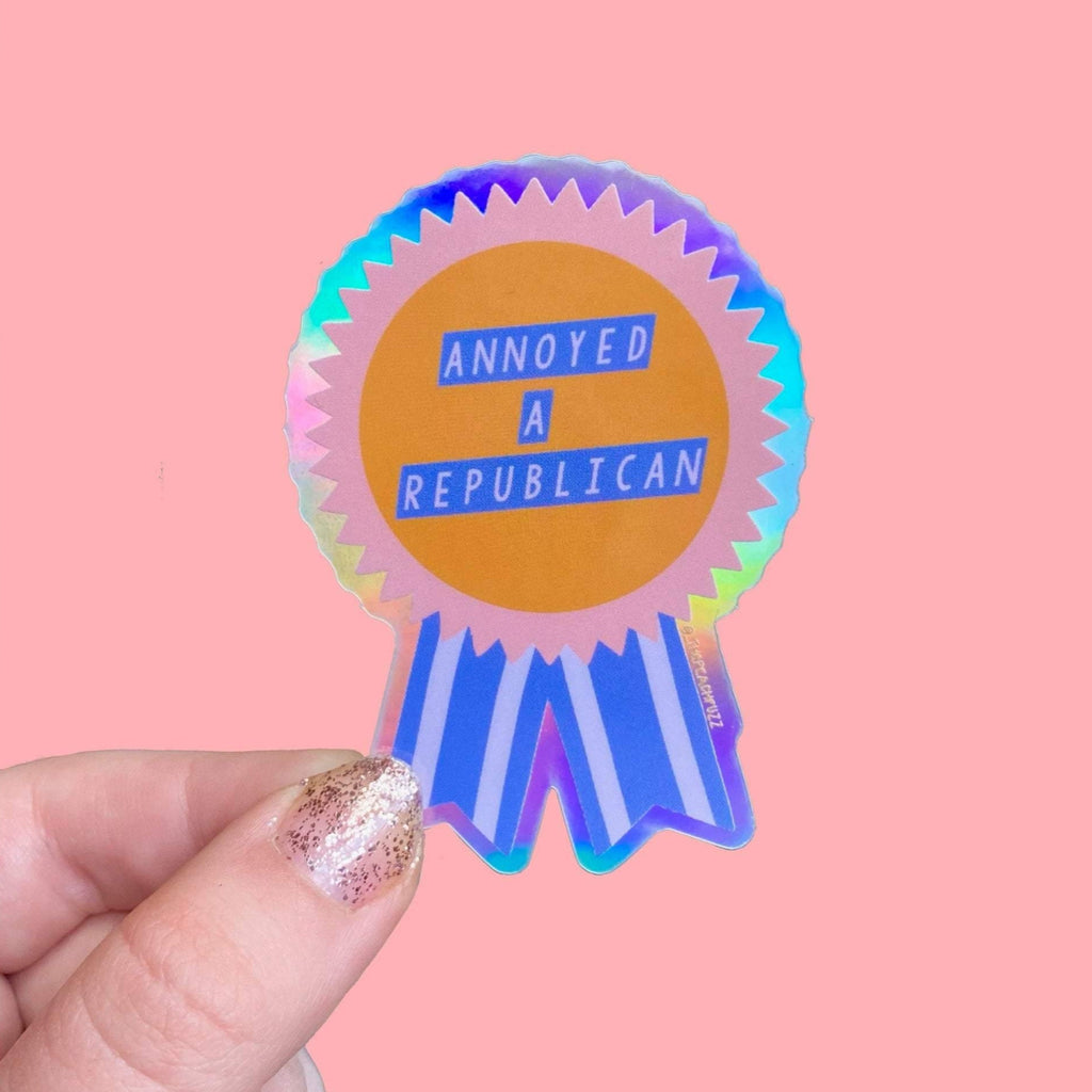 “Annoyed a Republican” Vinyl sticker with a glossy finish and a holographic outer border. Measures 1.9x2.7 inches.
