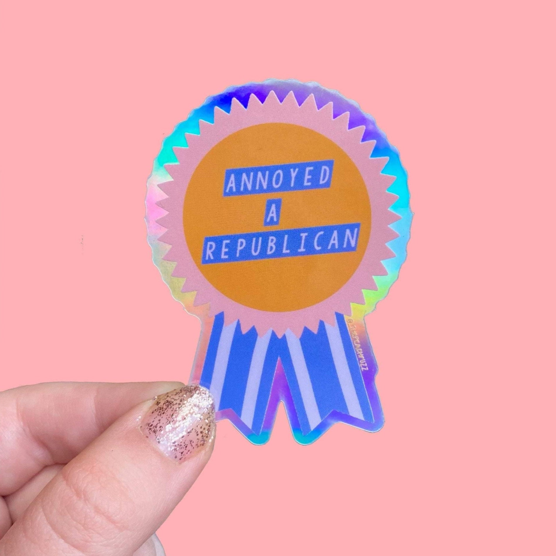 “Annoyed a Republican” Vinyl sticker with a glossy finish and a holographic outer border. Measures 1.9x2.7 inches.