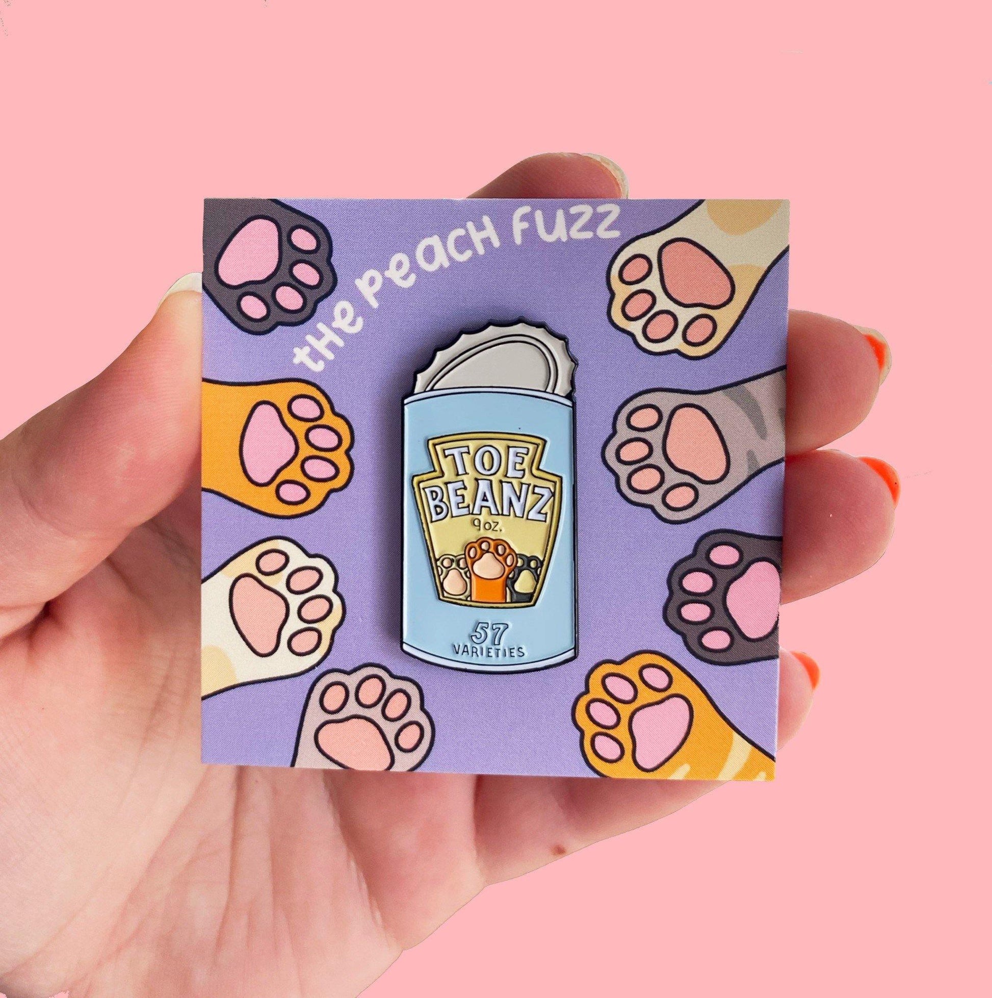 A soft enamel pin of a can of beans with the lid open and the label for the beans says “toe beans” with three little cat paws reaching up. The backing card has dark brown, yellow, white, orange, and gray cat paws reaching towards the center where the pin is.
