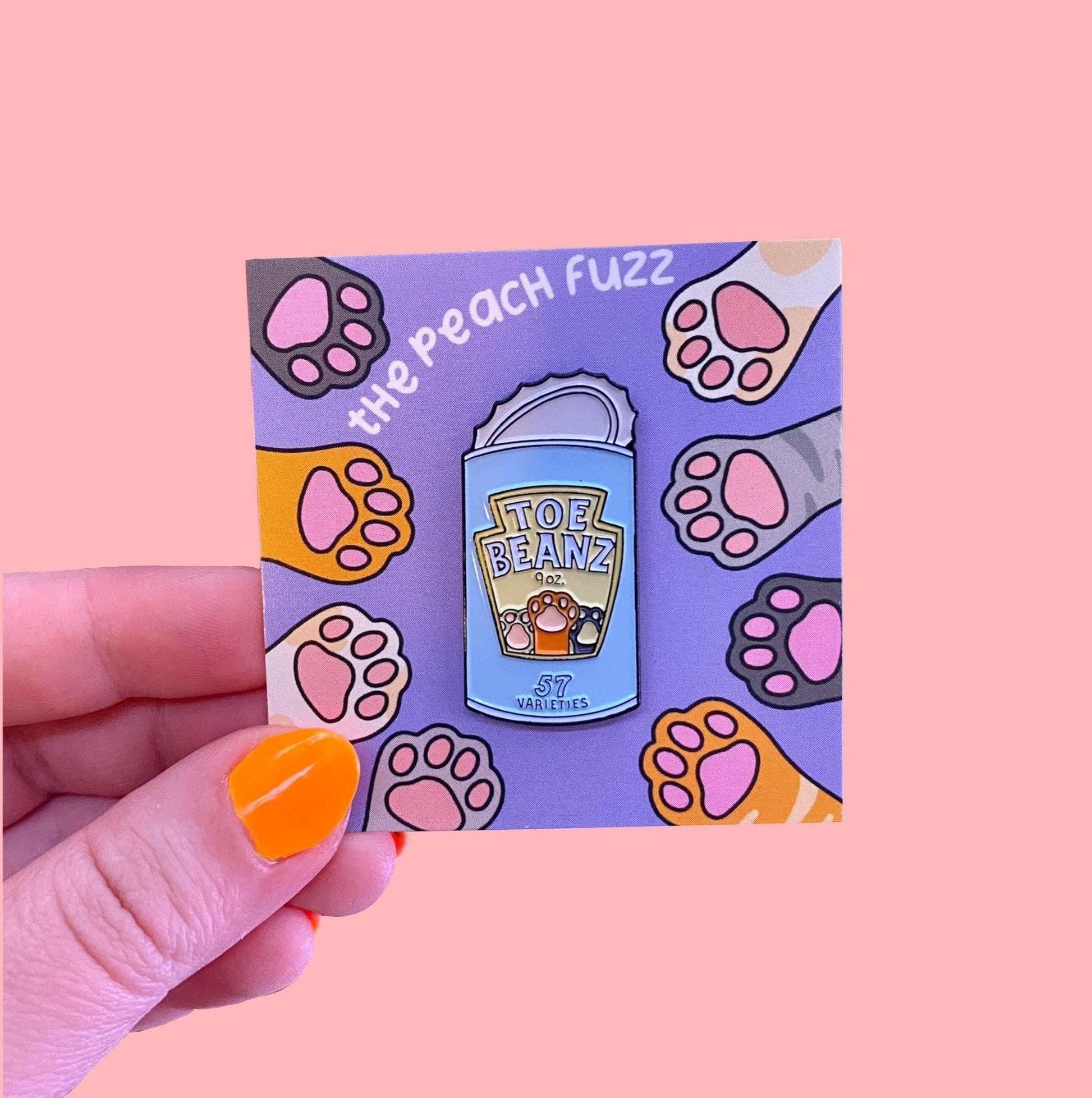 A soft enamel pin of a can of beans with the lid open and the label for the beans says “toe beans” with three little cat paws reaching up. The backing card has dark brown, yellow, white, orange, and gray cat paws reaching towards the center where the pin is.